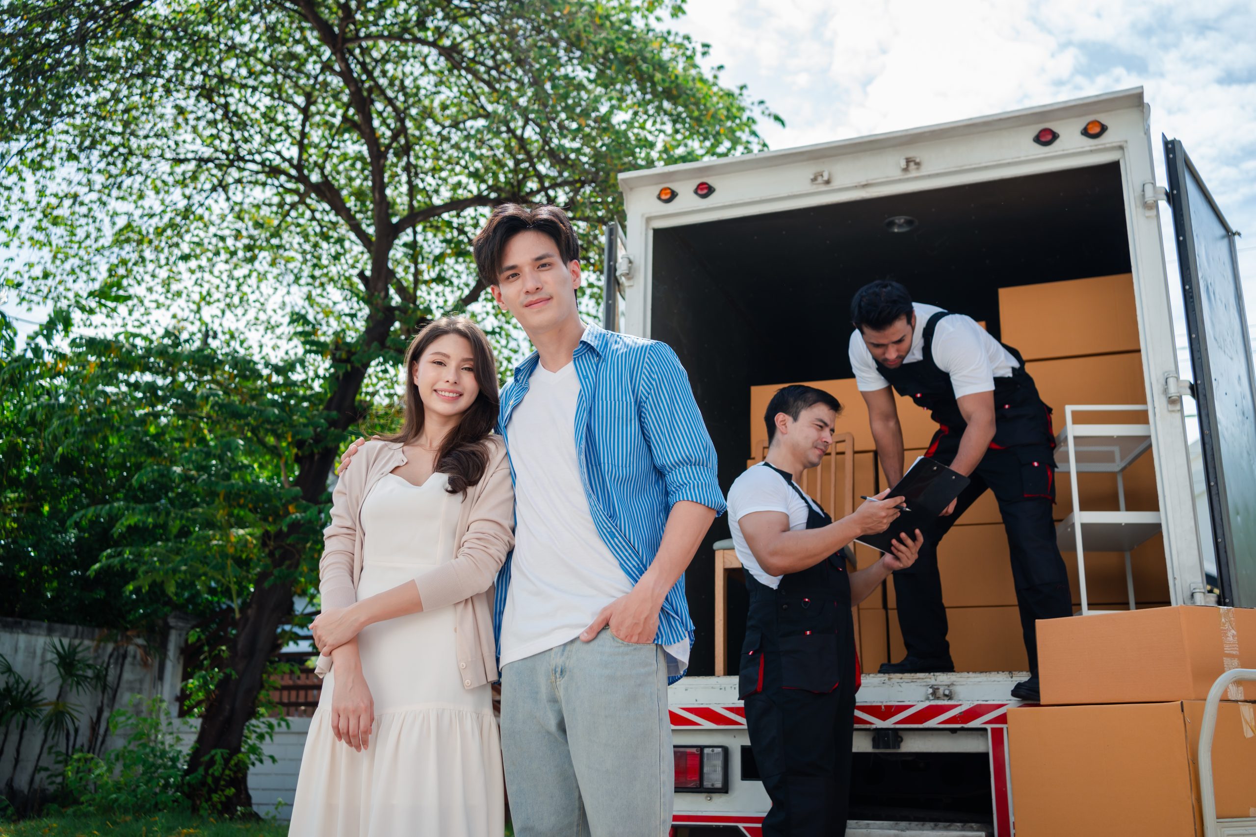 Asian Couple check while unloading boxes and furniture from a pickup truck to a new house with service cargo two men movers worker in uniform lifting boxes. concept of Home moving and delivery.